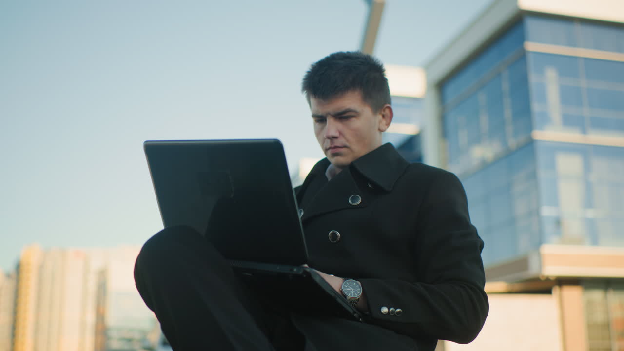 흑색 코트를 입은 부동산 거장, 도시 환경에서 노트북을 운영하는 데 집중하고, 손목에 눈에 띄는 손목 시계, 현대 사무실 건물과 배경의 도시 요소