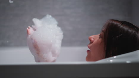 woman blows foam lying in bathtub at home. pleasured lady with soap bubbles on palms rests in bathroom. woman plays with shampoo froth in washroom