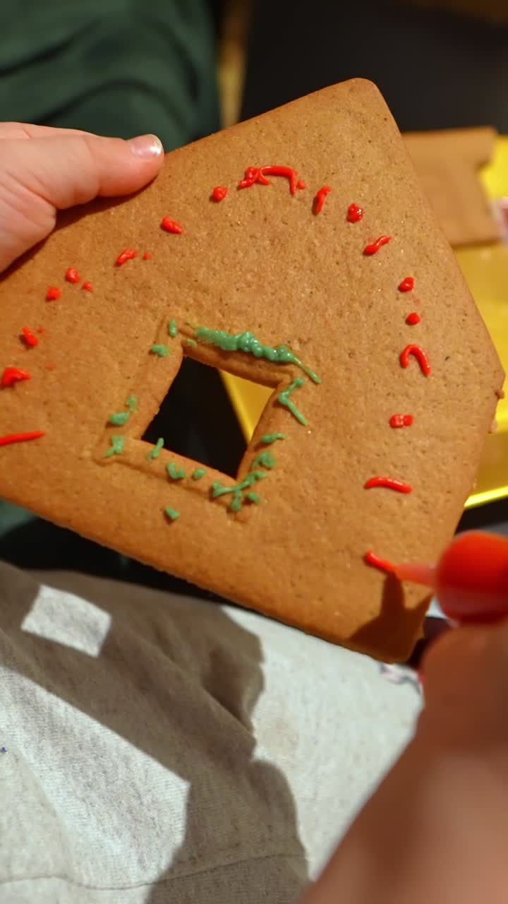 Young child decorates a gingerbread house piece with colorful frosting, close-up hands painting cookie icing during cozy Christmas baking, vertical shot