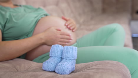 pregnant woman with bare belly at home. beautiful knitted blue baby socks on a sofa. future mother stroking her belly. pregnancy concept.