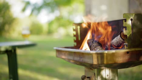 Fire-burning-for-a-barbecue-in-the-summer,-BBQ-getting-ready-in-a-green-garden-in-the-countryside,-with-cold-beers-on-a-picnic-table