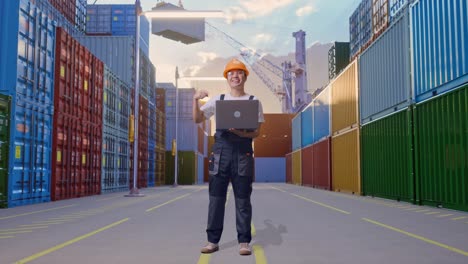 cuerpo lleno de hombre asiático trabajador con gafas de protección y casco de seguridad sonriendo y señalando a una computadora portátil en la mano mientras estaba de pie en el depósito de contenedores