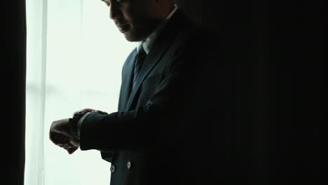 confident businessman in classic blue suit wearing or adjust watch with necktie near window in hotel room. handsome man wearing a nice suit on wedding day.