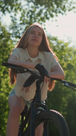 lower angle view of lady leaning on bike handlebar with a subtle smile, sunlight softly illuminating her face, background is filled with vibrant greenery