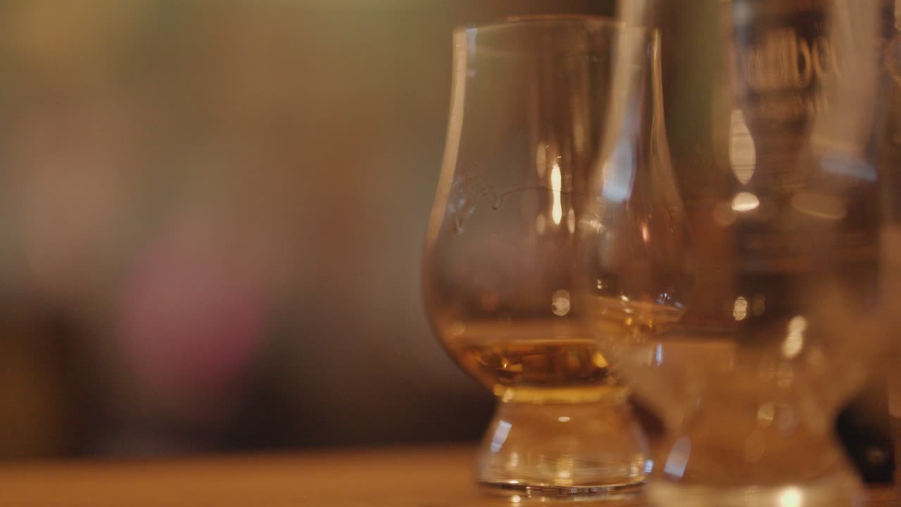 A hand selects a whiskey glass from a wooden bar counter in a warmly lit tasting room, with shallow depth of field and smooth camera focus
