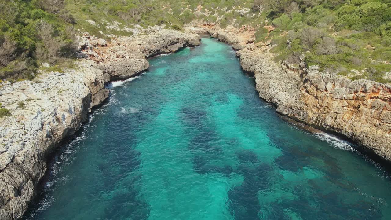 Flying with drone into a hidden cove surrounded by rugged rocky cliffs and dense green vegetation, filled with crystal-clear turquoise water.