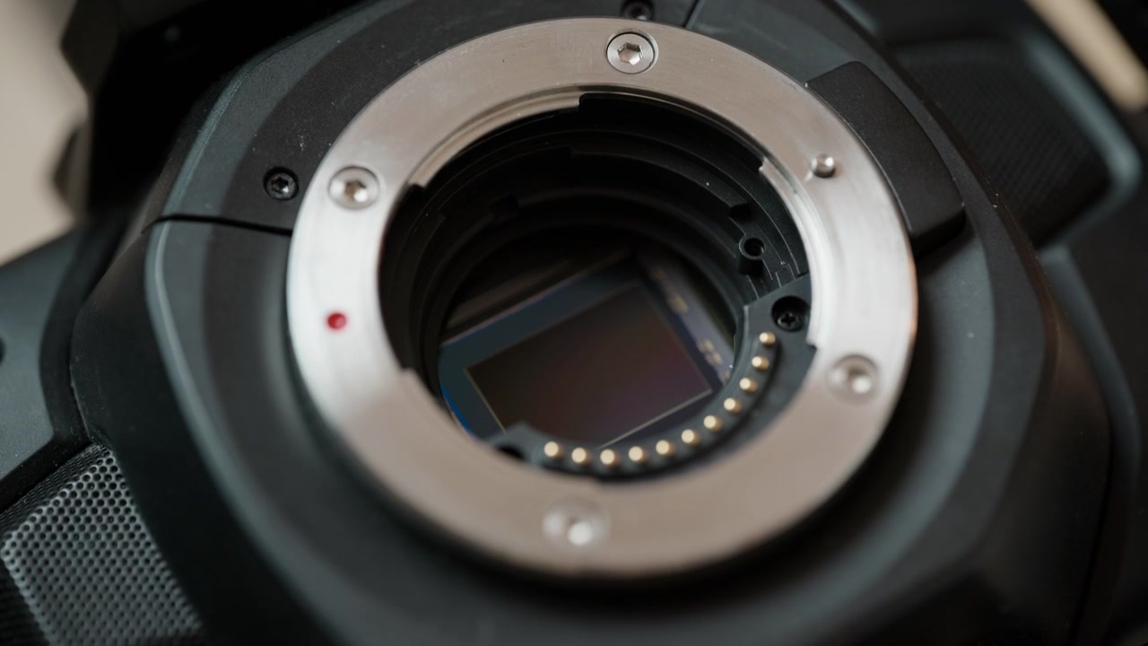 Close-up pinset mengangkat filter potong IR dari kamera sinema, memperlihatkan sensor gambar telanjang di baliknya. Langkah kunci modifikasi kamera spektrum penuh – pekerjaan sensor yang detail