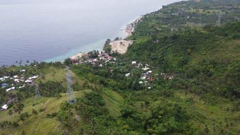 Electricity-lines-transmission-towers-above-seaside-village-between-tropical-green-coastal-foothill-terrain-on-Cebu-island,-Philippines