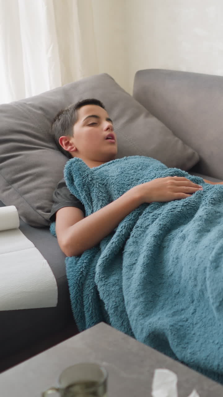Aerial view of scattered room with boy lying on couch under blanket, adjusting while glass cup and tissues are on the table, soft, cozy atmosphere with a sense of illness or rest
