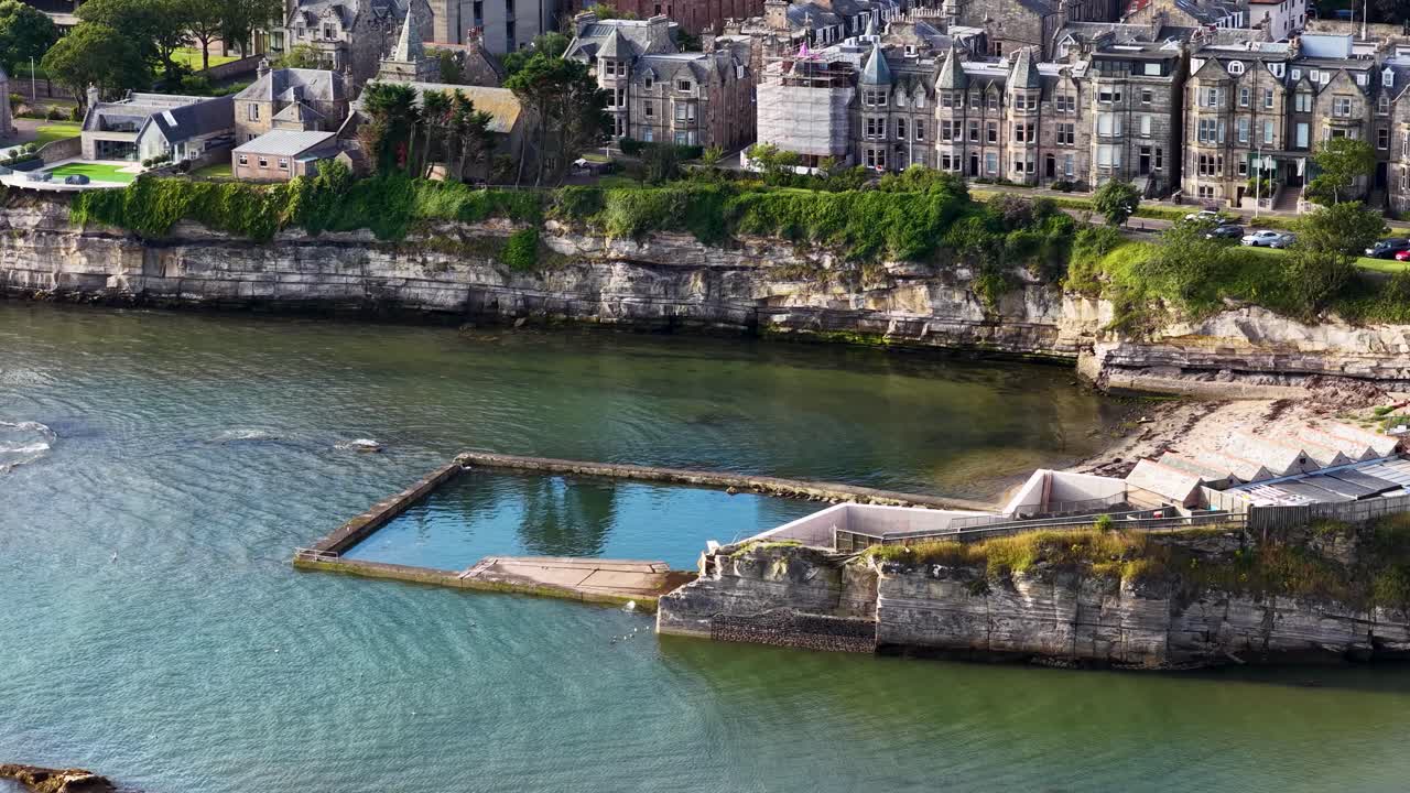 Drone slowly pans over tidal swimming pool, rocky coastline, and historic waterfront buildings in sunlight
