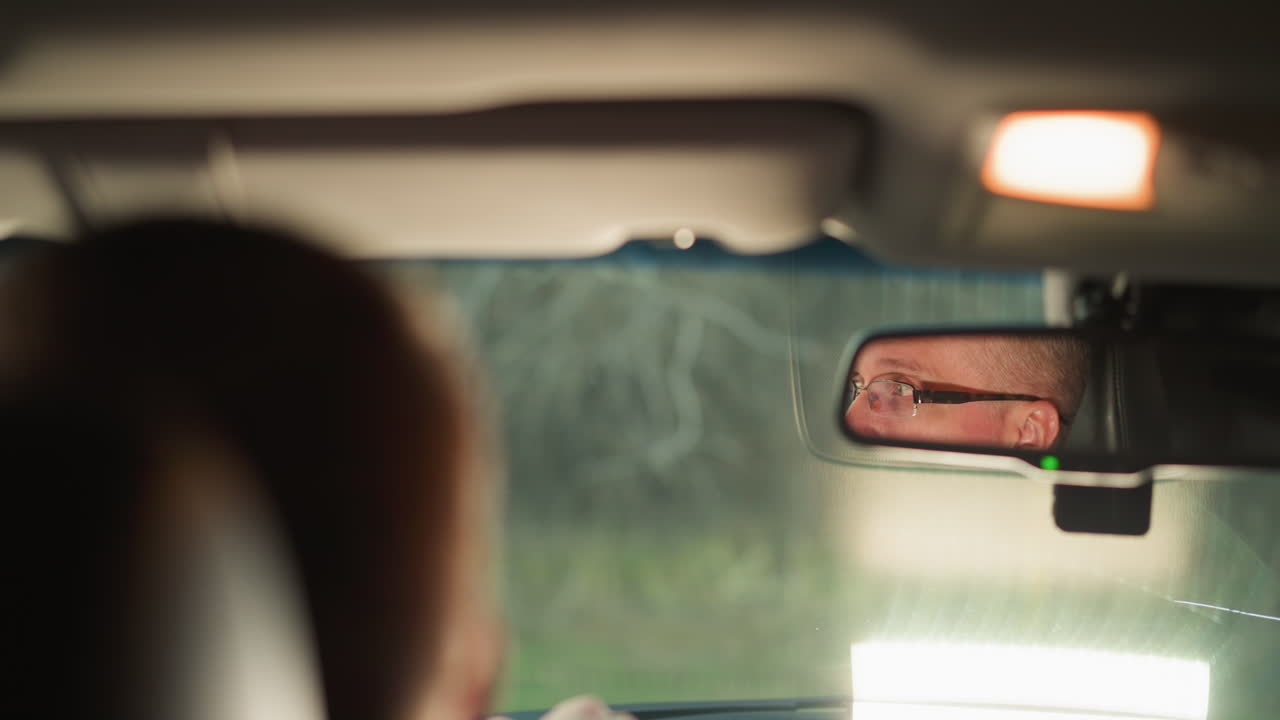 한 남자가 차 안에 앉아 있으며, 그의 얼굴은 뒷모습 거울에 명확하게 반사됩니다. 그의 표정은 집중적이고 약간 심각하며, 그는 집중하거나 깊은 생각에 빠져있다는 것을 암시합니다.