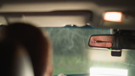 un hombre está sentado dentro de un coche, con su rostro reflejado claramente en el espejo retrovisor. su expresión está enfocada y ligeramente seria, lo que sugiere que está concentrado o profundamente pensado
