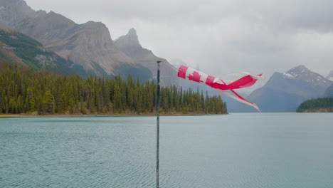 die kanadische flagge erwacht mit dem wind in der nähe eines atemberaubenden grünen wassersee, der von hohen bergen in den kanadischen rockies umgeben ist