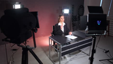caucasian lady wearing a black jacket sits at desk in studio. woman talks to camera in the light of soffits. high angle view.