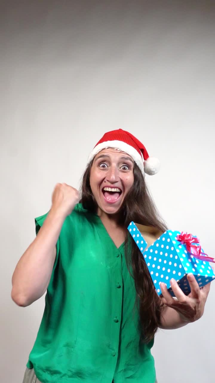 Joyful brunette in Santa hat celebrates blue polka-dot gift against white