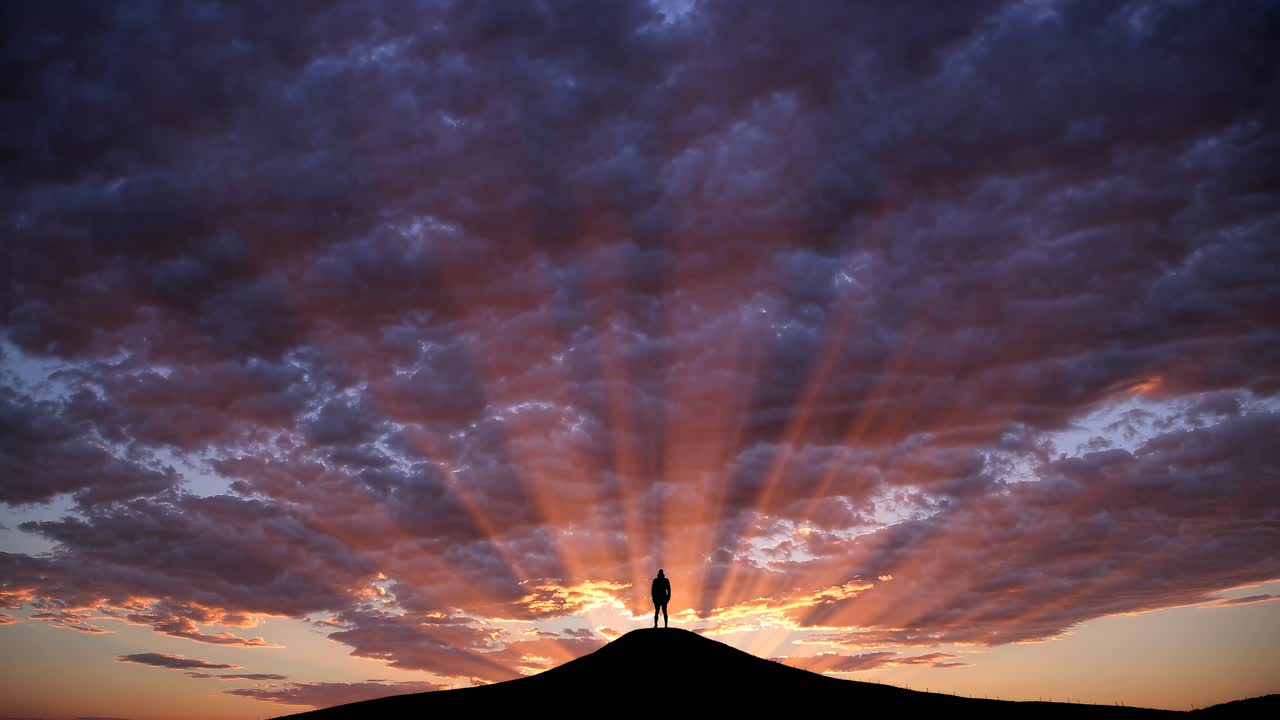 Silhouette of a person atop a hill at sunset, captured from a low-angle