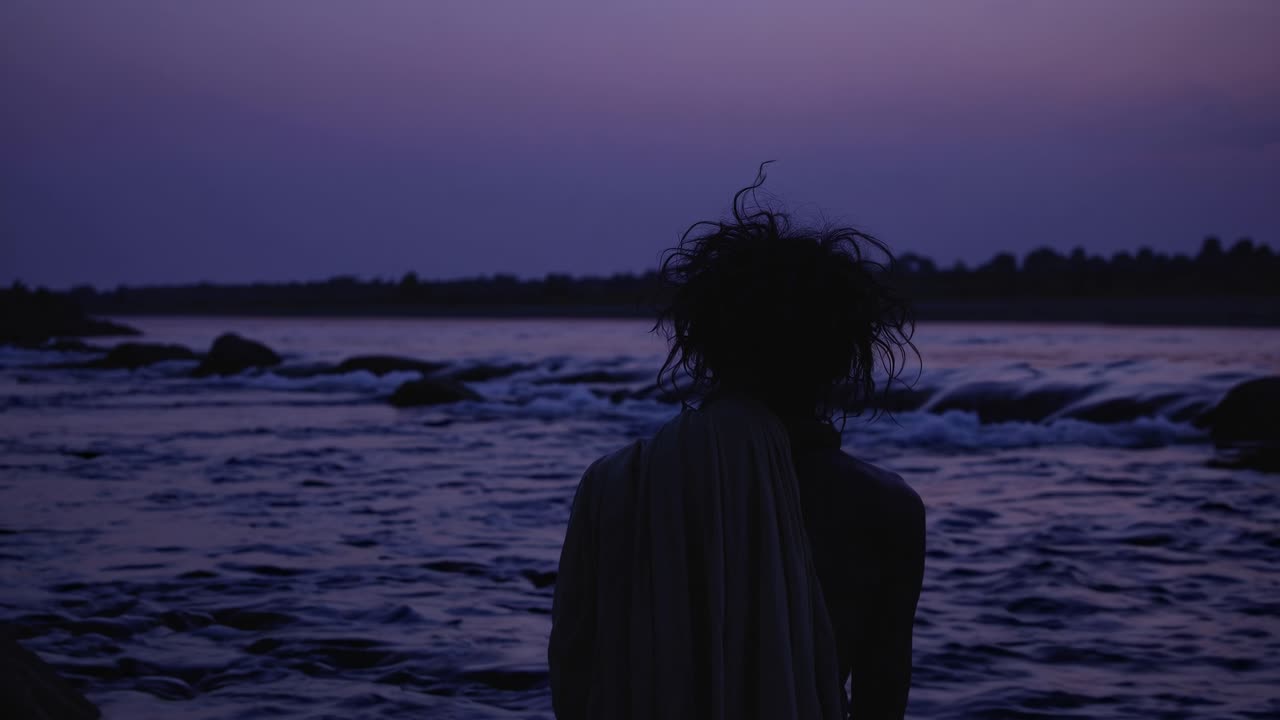 Rear view of a monk meditating near the river at twilight, enjoying the purple light of the sunset, creating a peaceful and spiritual atmosphere