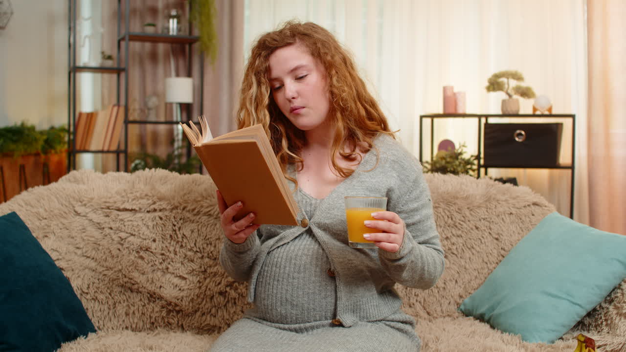 Pregnant young woman drinking orange juice while reading pregnancy book at home calm relaxed mood