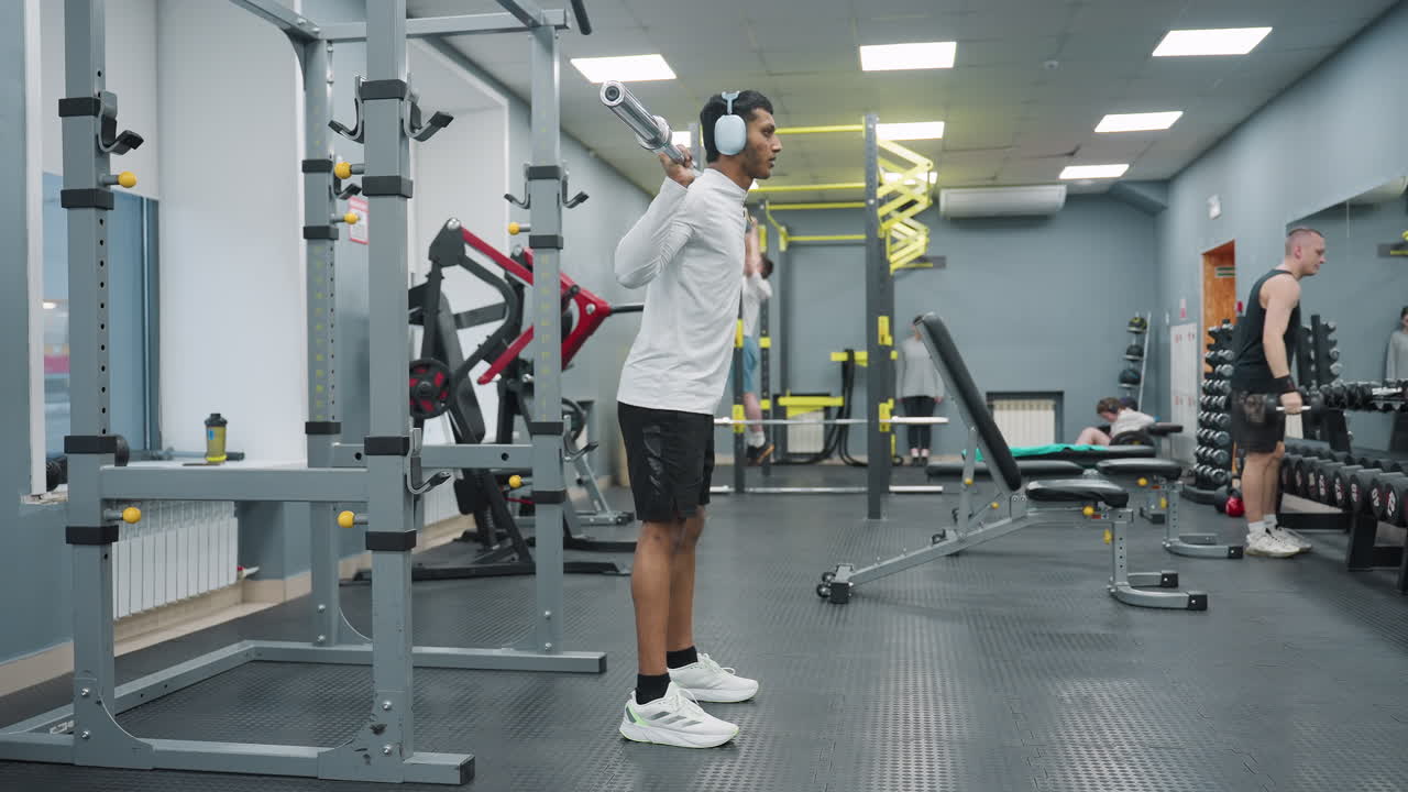 side view of gym rat squatting with weighted barbell while another person in background performs pull up on yellow workout rig in well equipped gym with strong lighting
