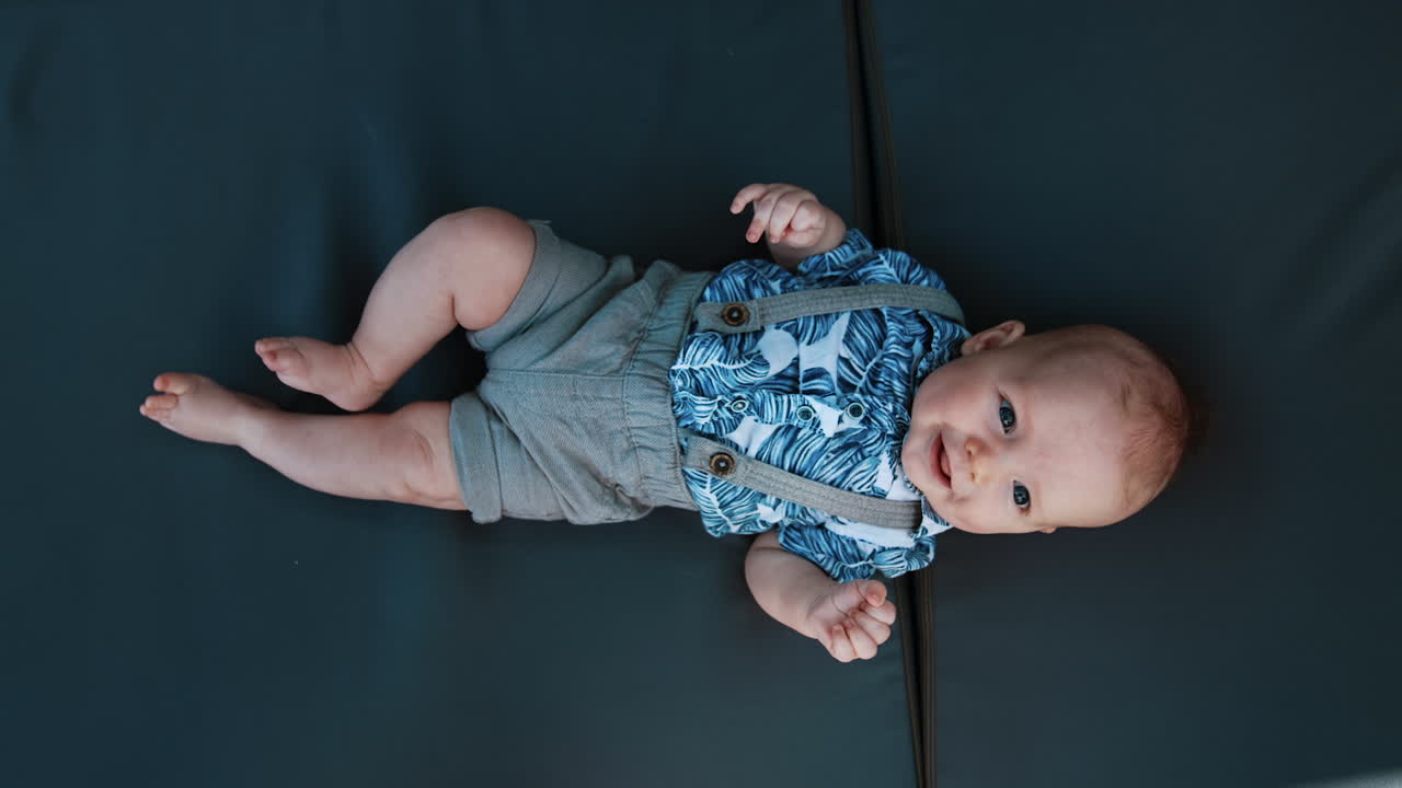 Adorable Caucasian baby boy wearing a blue shirt and shorts with suspenders. Smiling infant moving cheerfully his feet and hands. Vertical screen close up.
