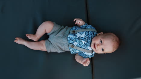 adorable caucasian baby boy wearing a blue shirt and shorts with suspenders. smiling infant moving cheerfully his feet and hands. vertical screen close up.