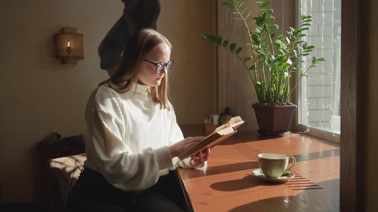 Real estate mogul sits quietly by window immersed in novel, flipping page as sunlight brightens tabletop with coffee mug, cozy seating, potted plant, and decorative interior details around her