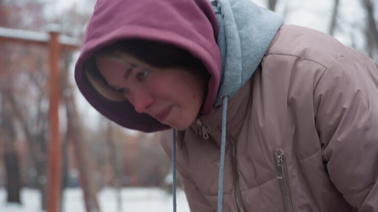 Una giovane donna in abbigliamento invernale si china in avanti, sentendo freddo, con uno sfondo innevato sfocato e alberi spogli. Il cappuccio della giacca le copre la testa, e il clima rigido è evidente nell'ambiente invernale esterno.