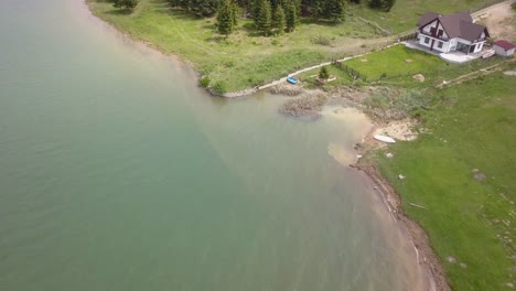 aerial panning shot of lake meeting woodland land with white house in the frame