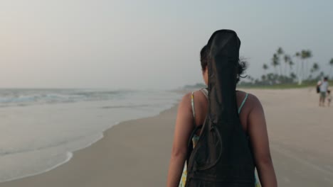 Young-woman-with-guitar-case-walking-on-beach-at-dusk,-palm-trees-in-background,-couple-in-distance