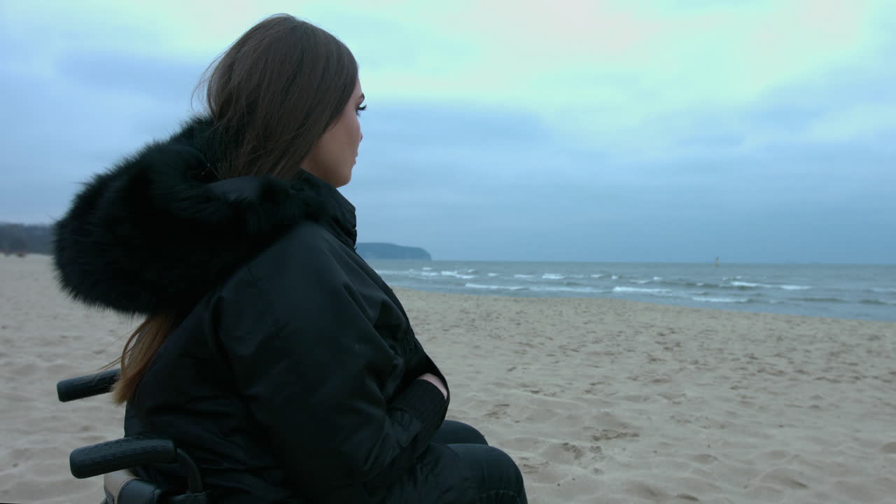 A disabled woman in a wheelchair alone at the beach observing around. A woman sitting in a wheelchair at the beach watching the waves in a thoughtful moment. A disabled woman in a relaxing expression
