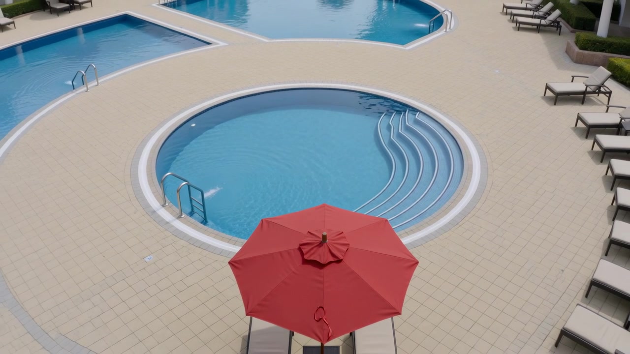 Drone trækker sig tilbage og afslører en rund pool, rød parasol på dækket for at vise indretningen, med to gæster.