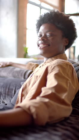 vertical video of portrait of happy african american man sitting on sofa at home, slow motion