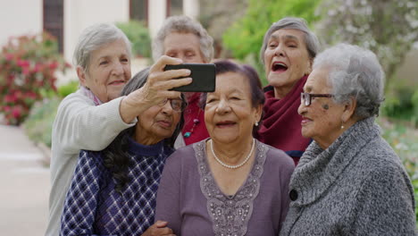Eine-Gruppe-Multiethnischer-älterer-Frauen-Posiert-Mit-Einem-Smartphone-Und-Macht-Lächelnd-Ein-Selfie-Foto.-Sie-Genießen-Einen-Glücklichen,-Unbeschwerten-Lebensstil-Im-Ruhestand-Im-Schönen-Garten-Im-Freien-Und-Teilen-Erinnerungen