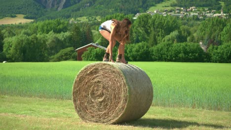 a beautiful brunette woman standing up and balancing carefully on a large bale of hay on a farm in france