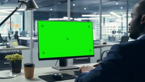 empresario negro exitoso sentado en un escritorio trabajando en una computadora portátil de pantalla verde en la oficina. empresario afroamericano usando una pantalla de llave cromada. disparo estático sobre el hombro