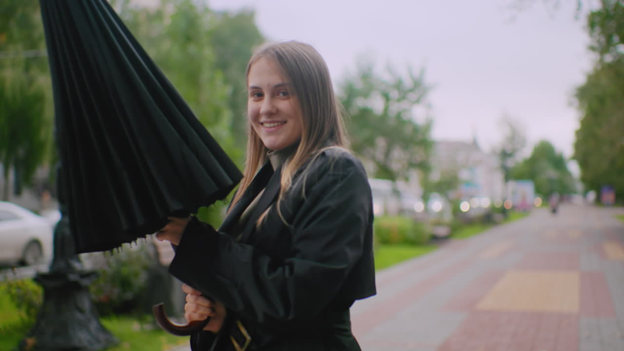 미소를 지으며 검은 우산을 접는 여성의 모습이에요. 흐린 날씨에 자동차, 건물, 나무, 사람들이 흐릿하게 보이는 도시 인도에 서 있는 모습에서 도시적인 분위기와 라이프스타일을 느낄 수 있어요.