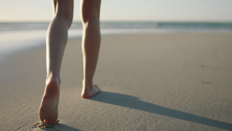 Cerca-De-Las-Piernas-De-Una-Mujer-Caminando-Sobre-Los-Tranquilos-Pies-De-La-Playa-Junto-Al-Mar-Dejando-Huellas-En-La-Arena