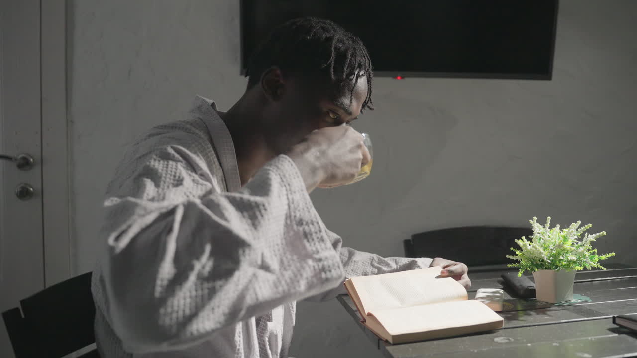 Champ reading book at table, holding glass of tea, relaxed morning in cozy home environment with sunlight, potted plant in background, peaceful, calm atmosphere