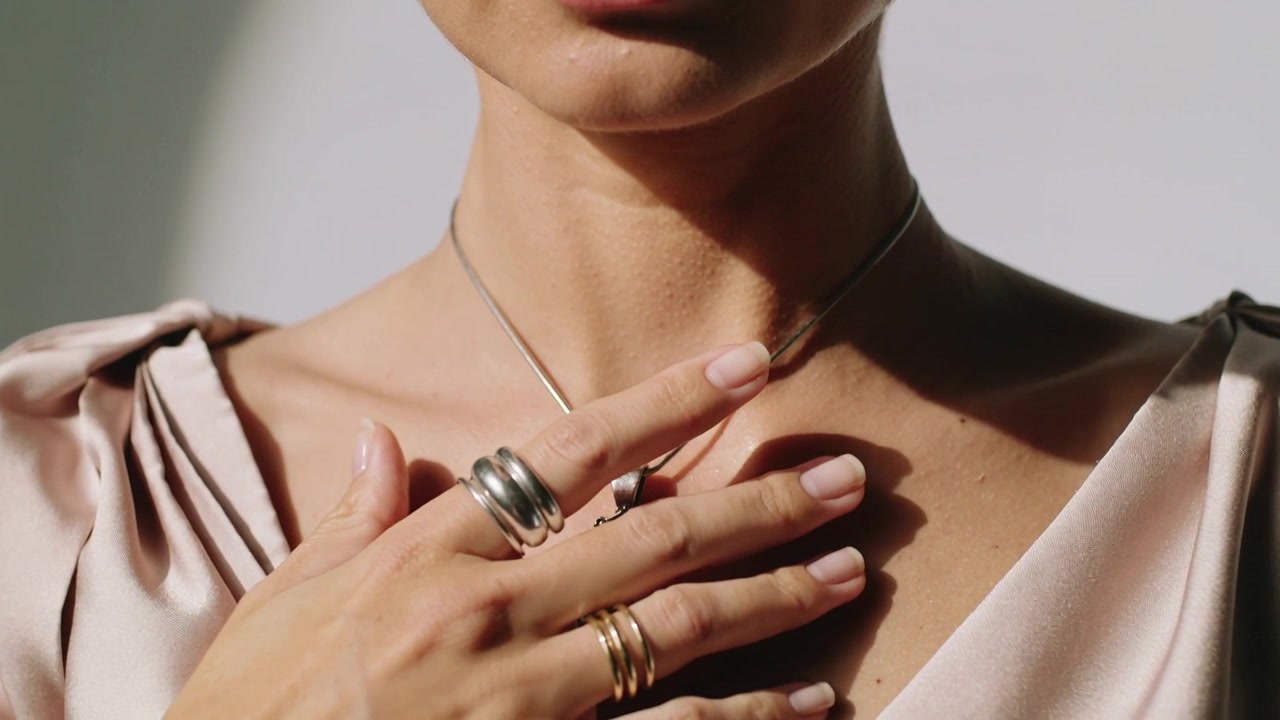 Donna in studio che alza la mano destra per sistemare un ciondolo d'argento al collo, con anelli e un top di raso.