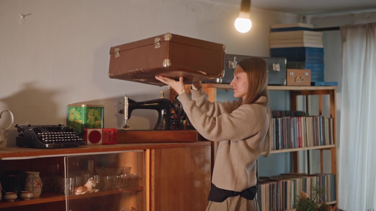 Mujer caucásica alcanzando una maleta en el estante superior, levantando un baúl de cuero de un estante alto bajo el cálido resplandor de una lámpara, con una colección de discos de fondo, escena de evocación de recuerdos de viaje, delicado esfuerzo y expectación.