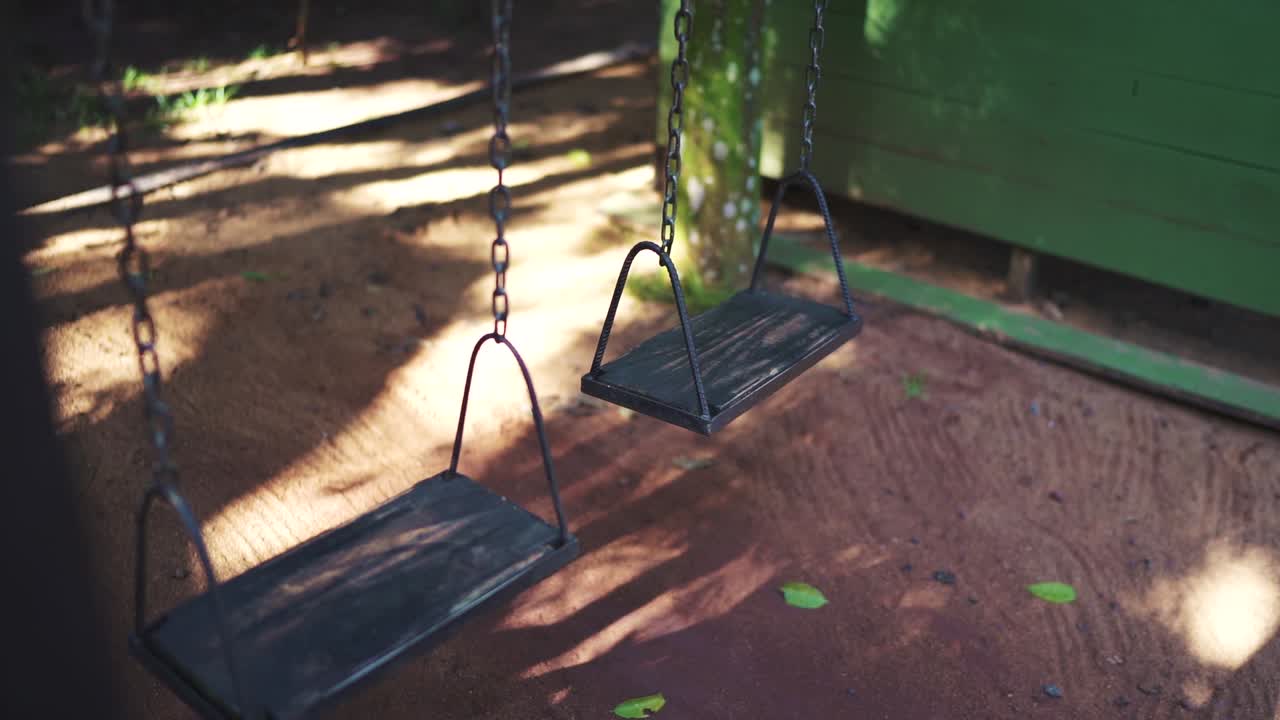 Due altalene vuote pendono in un parco giochi tranquillo, proiettando ombre delicate sul terreno illuminato dal sole, creando un'atmosfera serena e nostalgica.