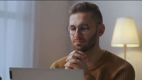 Un-Joven-Con-Gafas-Está-Escuchando-Y-Viendo-Una-Conferencia-O-Un-Discurso-De-Un-Participante-De-Una-Reunión-En-Línea-Sentado-En-La-Oficina-De-Su-Casa,-Comunicación-A-Distancia-Y-Autoeducación.