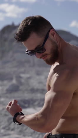 handsome bodybuilder on the white hill. strong athlete without shirt shows his arm biceps on nature background. healthy and sport lifestyle. slow motion. vertical video
