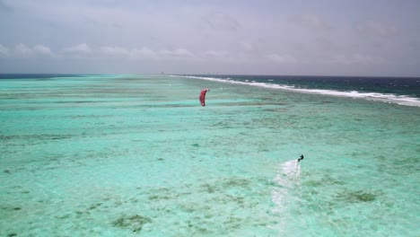 Un-Kitesurfista-Solitario-Deslizándose-Sobre-Las-Vibrantes-Aguas-Turquesas-De-Los-Roques-Con-Una-Barrera-De-Coral-Distante,-Vista-Aérea