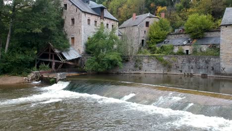 River-pouring-over-dam-at-the-foot-of-old-farm-buildings,-drone-shot