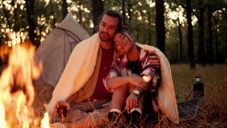 a brunette guy and a black-skinned girl are sitting and hugging under a white blanket near a fire against the backdrop of a tent in a green forest during a hike. happy and cozy evening during a rest in the evening during a hike