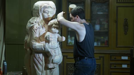 a sculptor in a sleeveless shirt uses a chisel and mallet to carve intricate details into a large, unfinished wooden madonna and child statue in a craftsman’s studio