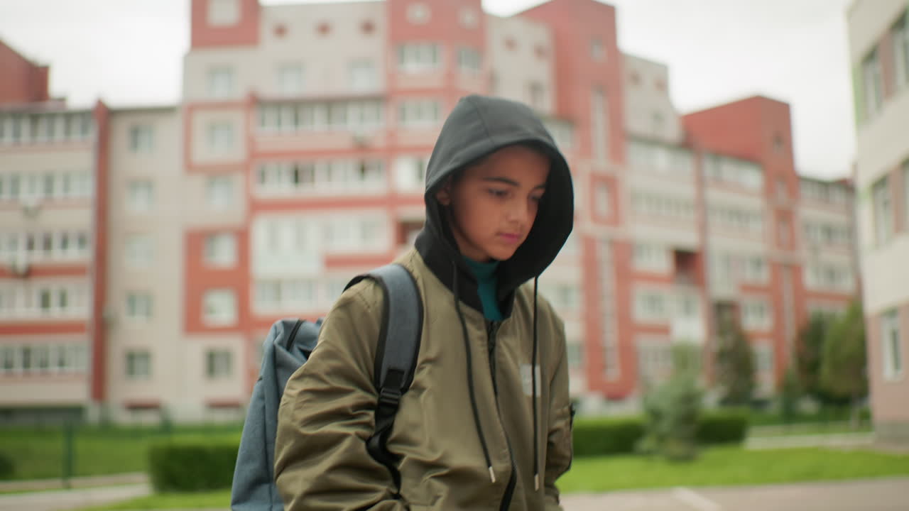 niño con sudadera con capucha y mochila caminando hacia el aula con una sonrisa cálida, fondo borroso que muestra una figura distante y altos edificios residenciales alrededor del campus