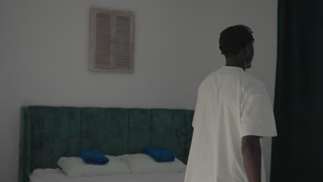 man wearing white t-shirt bends over in minimalist bedroom, focusing on something near the bed, with sunlight streaming in, simple room design with green headboard and dark wood door visible