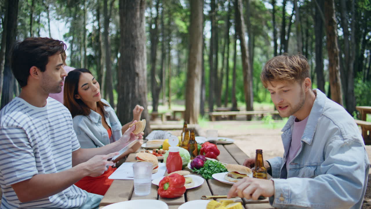 Young people eating picnic table in summer forest. Happy men women enjoy meal